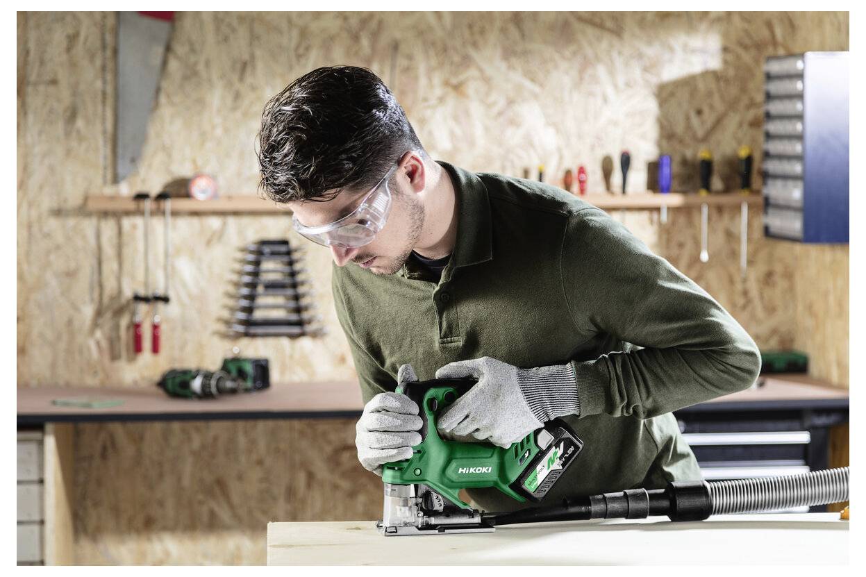 Ein Mann mit Sicherheitsschutzbrille verwendet eine grüne Kreissäge an einem Holzbrett in einer Werkstatt und zeigt konzentrierte Aufmerksamkeit beim Schneiden.