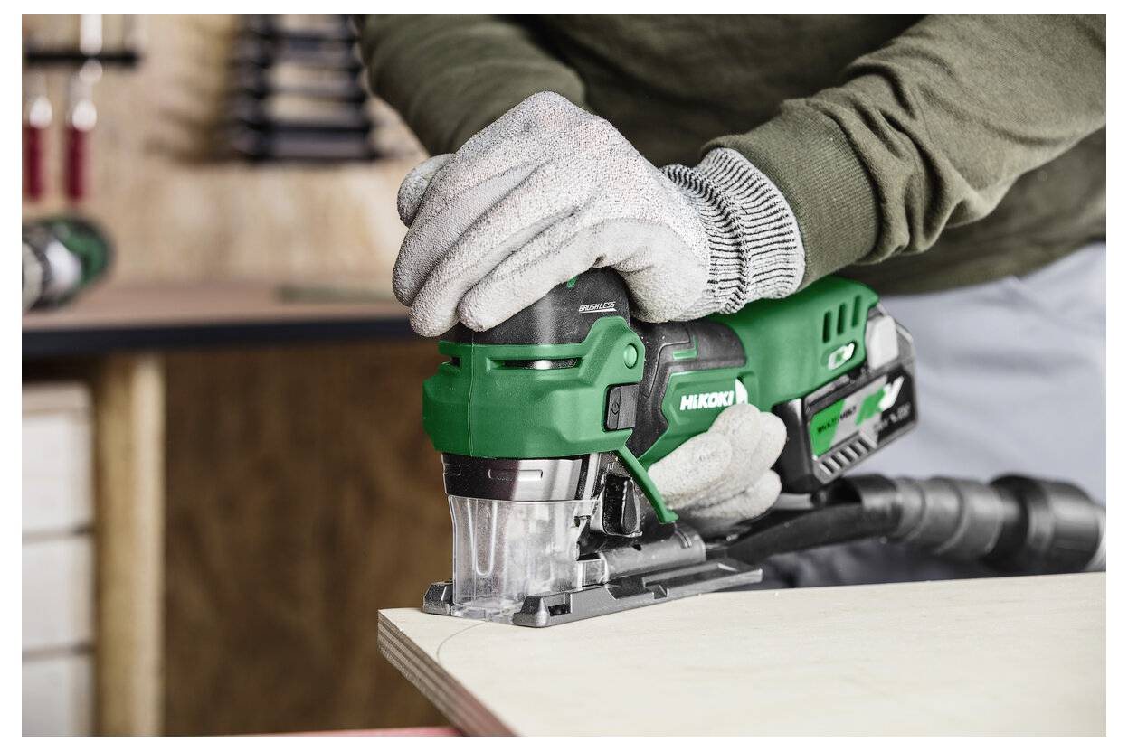 Eine Person mit Handschuhen benutzt eine grüne elektrische Stichsäge, um ein Stück Holz zu schneiden. Werkzeuge sind im unscharfen Hintergrund zu sehen.
