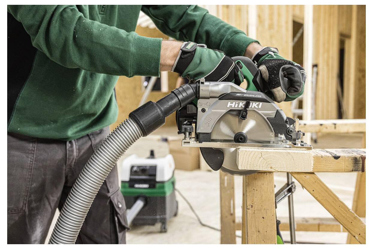 Eine Person, die mit einer Kreissäge Holz schneidet und dabei grüne Handschuhe und ein grünes Oberteil trägt, in einer Werkstattumgebung mit weiteren Werkzeugen im Hintergrund.