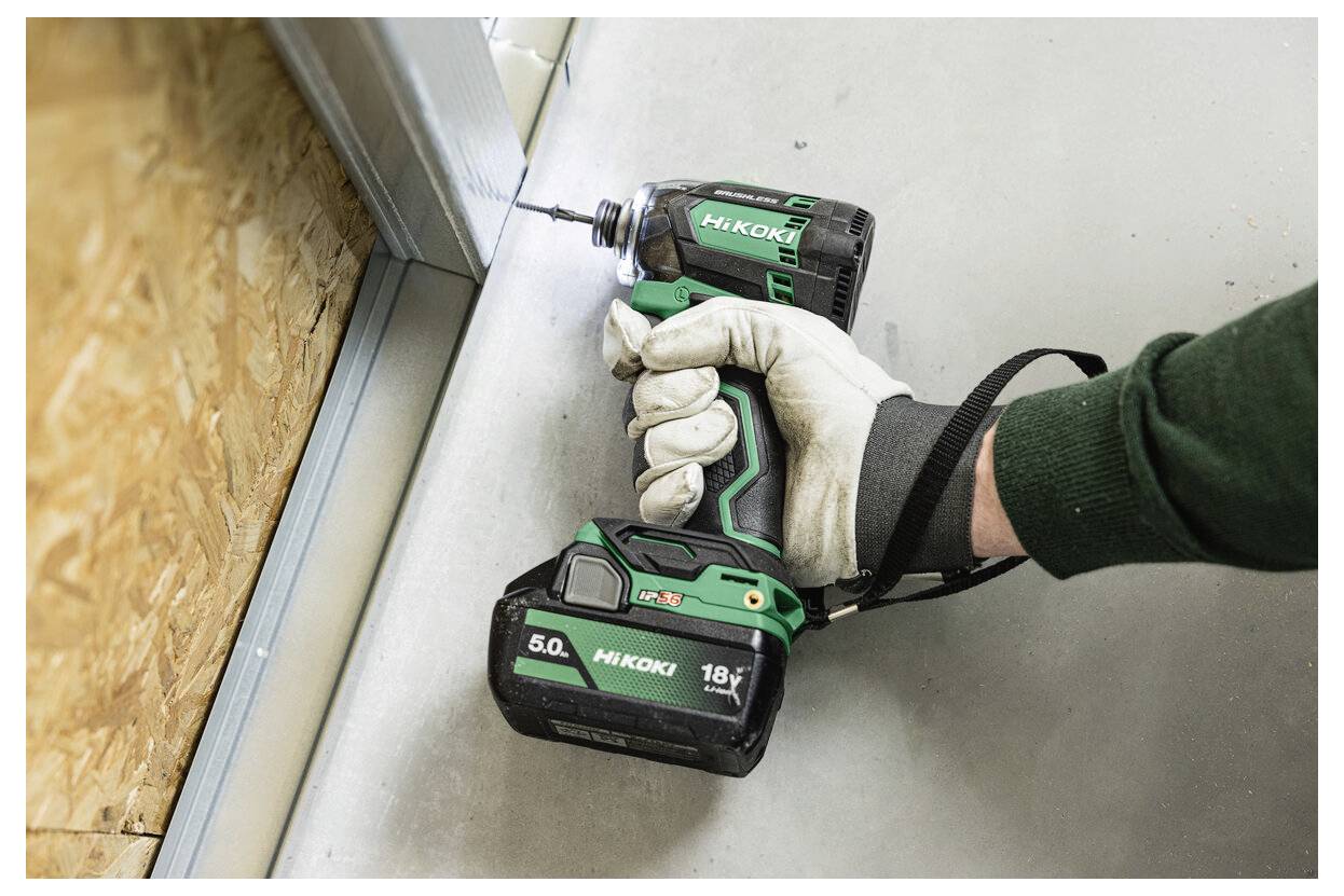 Eine Person mit Handschuhen benutzt einen grünen Akkuschrauber, um einen Metallrahmen auf einem Betonboden neben einer Holzwand zu befestigen.