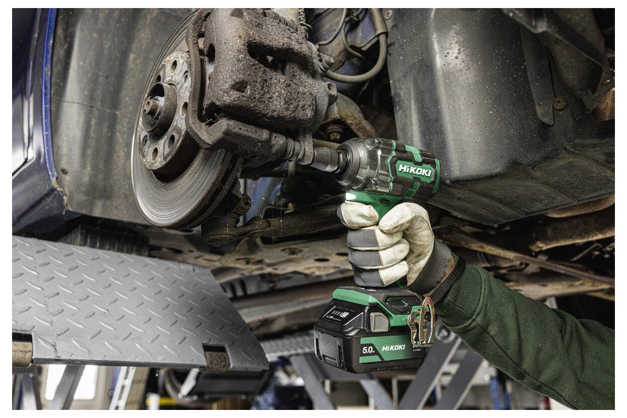 Eine Handschuh tragende Hand bedient eine kabellose Schlagschrauber an der Bremsanlage eines Autos in einer Garage. Das Werkzeug ist grün und schwarz und wird fest gehalten.