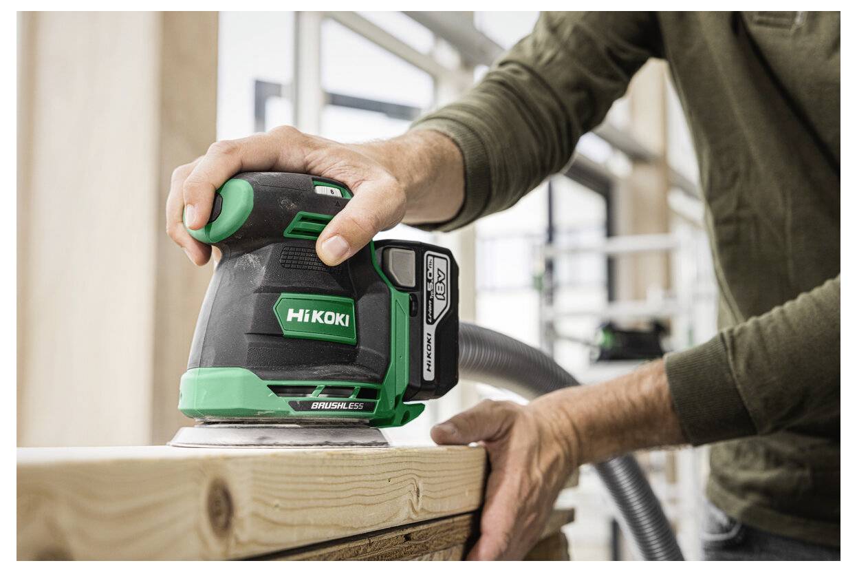 Eine Person verwendet einen grün-schwarzen Schleifer auf einer Holzoberfläche in einer Werkstatt und glättet die Kante eines hellfarbenen Holzbretts.