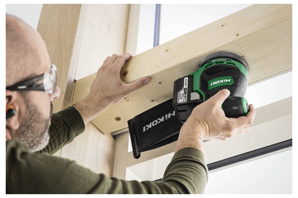 Eine Person trägt Schutzbrille und Gehörschutz und verwendet einen grünen Hitachi-Schleifer an einem Holzbalken in Innenräumen.