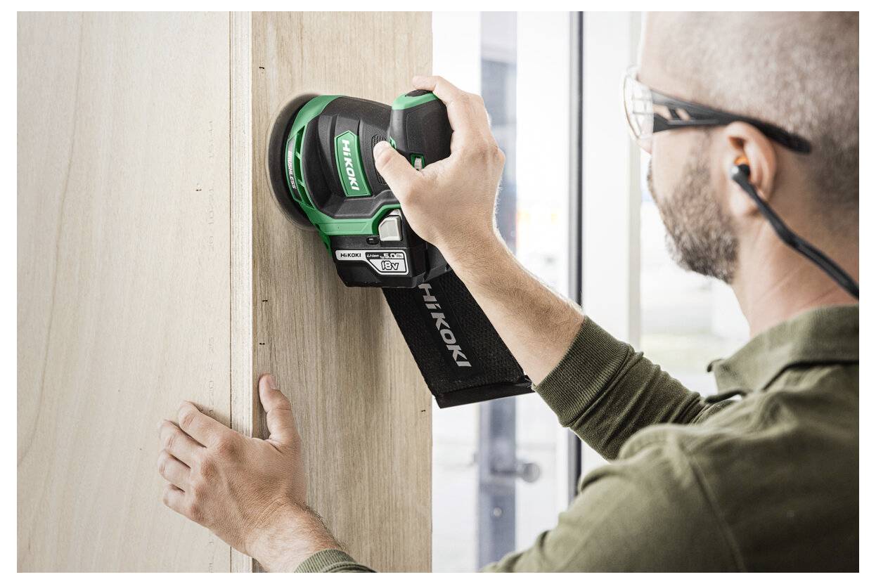 Eine Person, die einen grün-schwarzen Elektro-Schleifer auf einer Holzoberfläche verwendet, mit Schutzbrille und Gehörschutz, in Richtung eines Fensters.