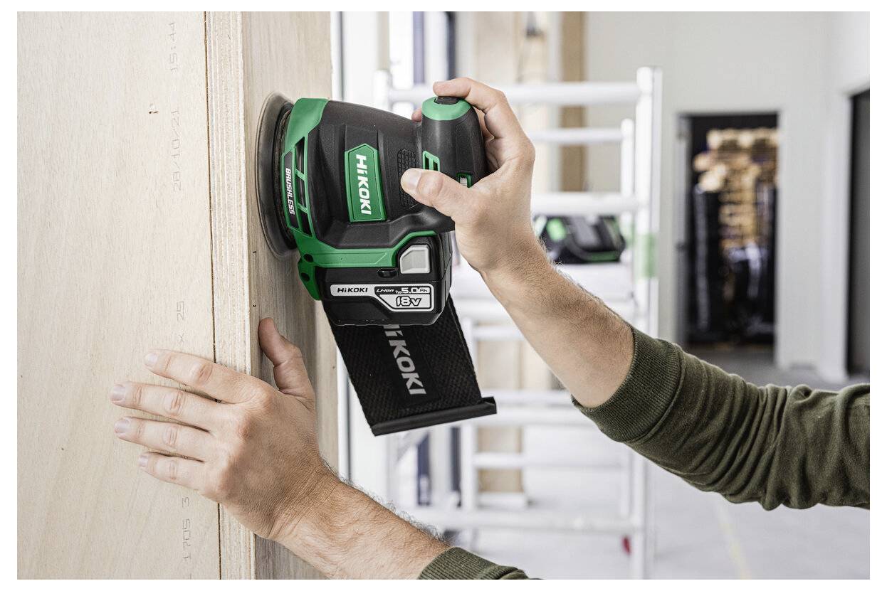 Eine Person verwendet einen grünen HiKOKI-Elektroschleifer, um eine Holzoberfläche in einer Werkstatt zu glätten, wobei Werkzeuge im Hintergrund zu sehen sind.