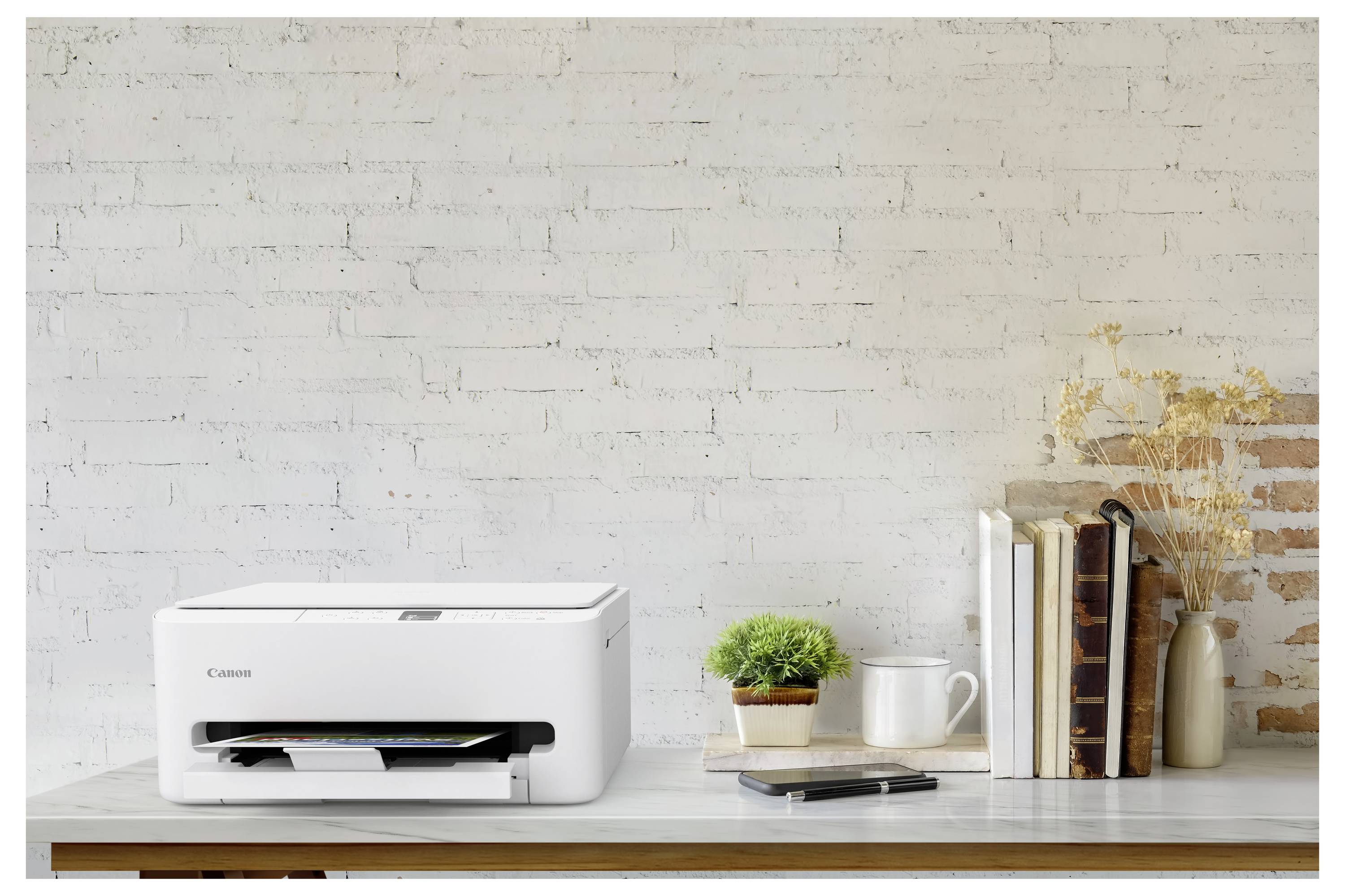 Ein kompakter weißer Drucker auf einem Holzschreibtisch neben einer kleinen Pflanze, Büchern und einem Smartphone, vor einer weißen Ziegelwand im Hintergrund.