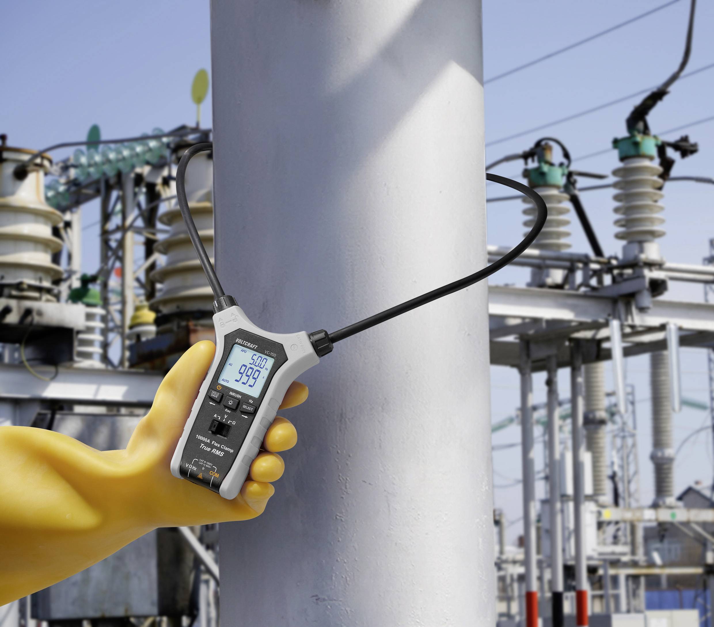 Eine Hand in einem gelben Handschuh hält einen digitalen Zangenstrommesser, der '999' anzeigt und den Strom um einen Pfosten in einem industriellen Kraftwerk misst.