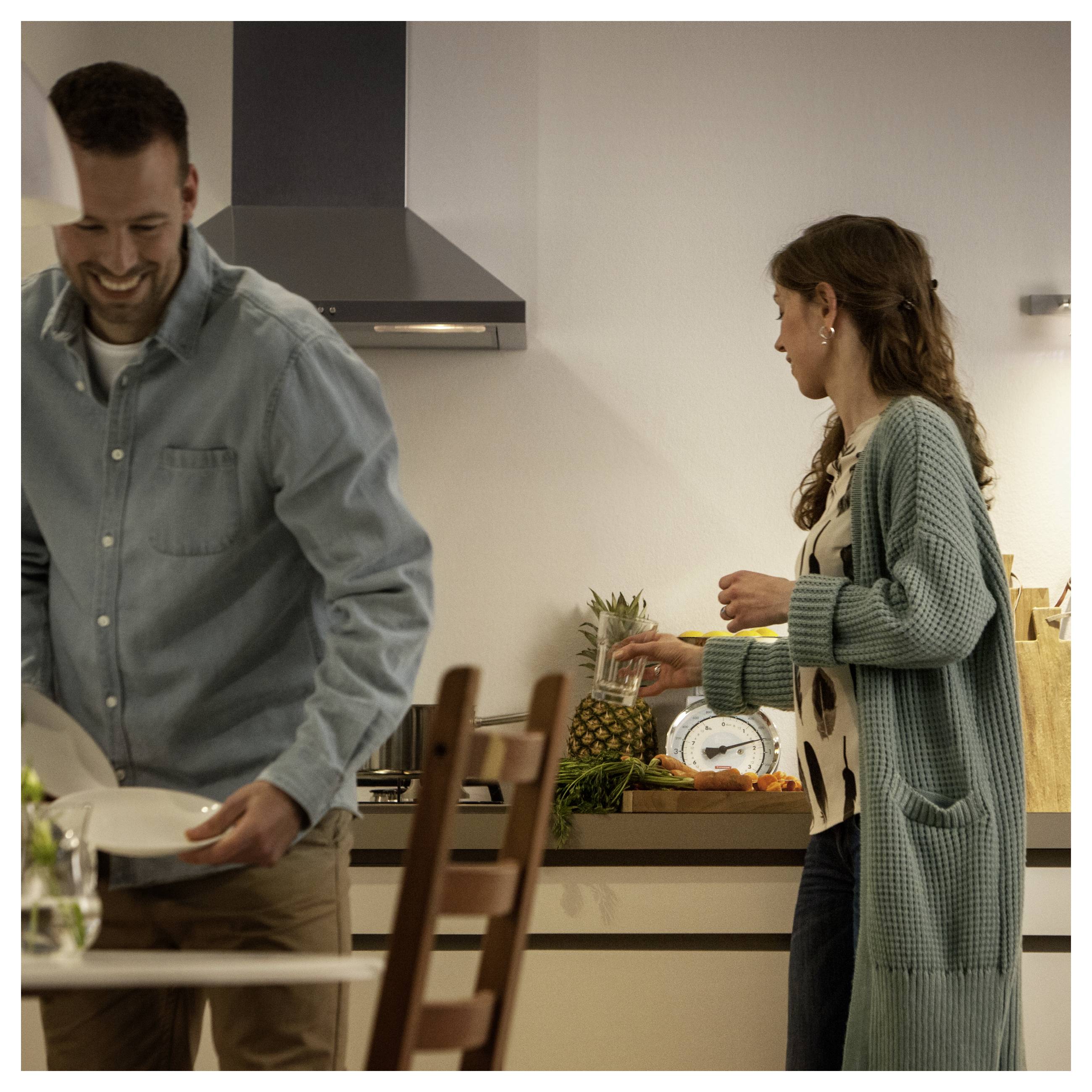 Ein Mann und eine Frau in einer Küche; sie schneidet Gemüse und eine Ananas, während er den Tisch deckt, beide lächeln und unterhalten sich.