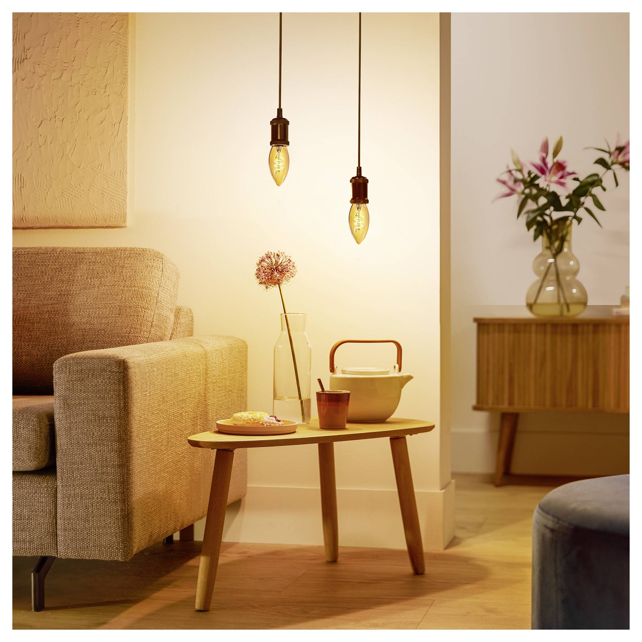 Eine gemütliche Ecke im Wohnzimmer mit einem beigen Sessel, einem Holztisch, einer Vase mit rosa Blumen und zwei hängenden Glühbirnen, die eine warme Atmosphäre schaffen.