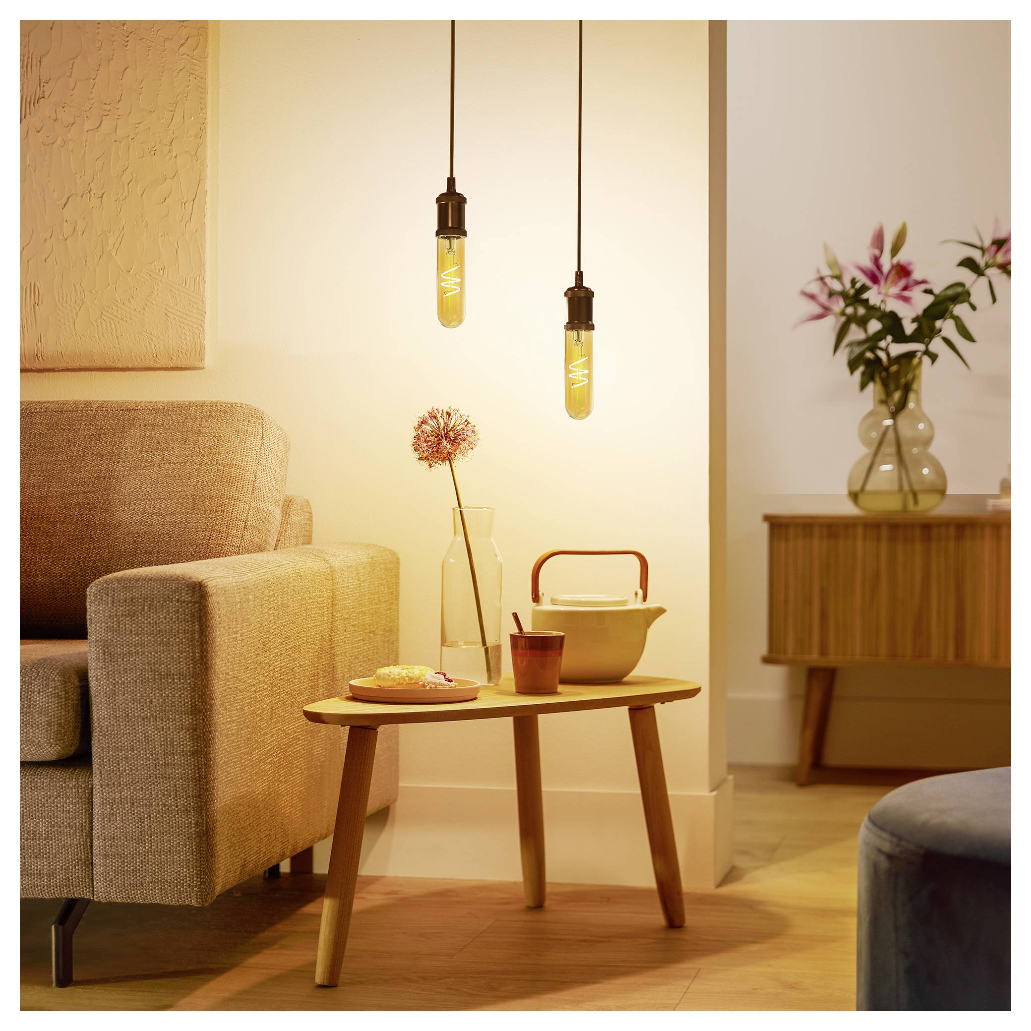 Eine gemütliche Ecke des Wohnzimmers mit einem beigen Sofa, hängenden Edison-Glühbirnen, einem Holzcouchtisch mit einer pinken Blume in einer Vase und einem Sideboard.