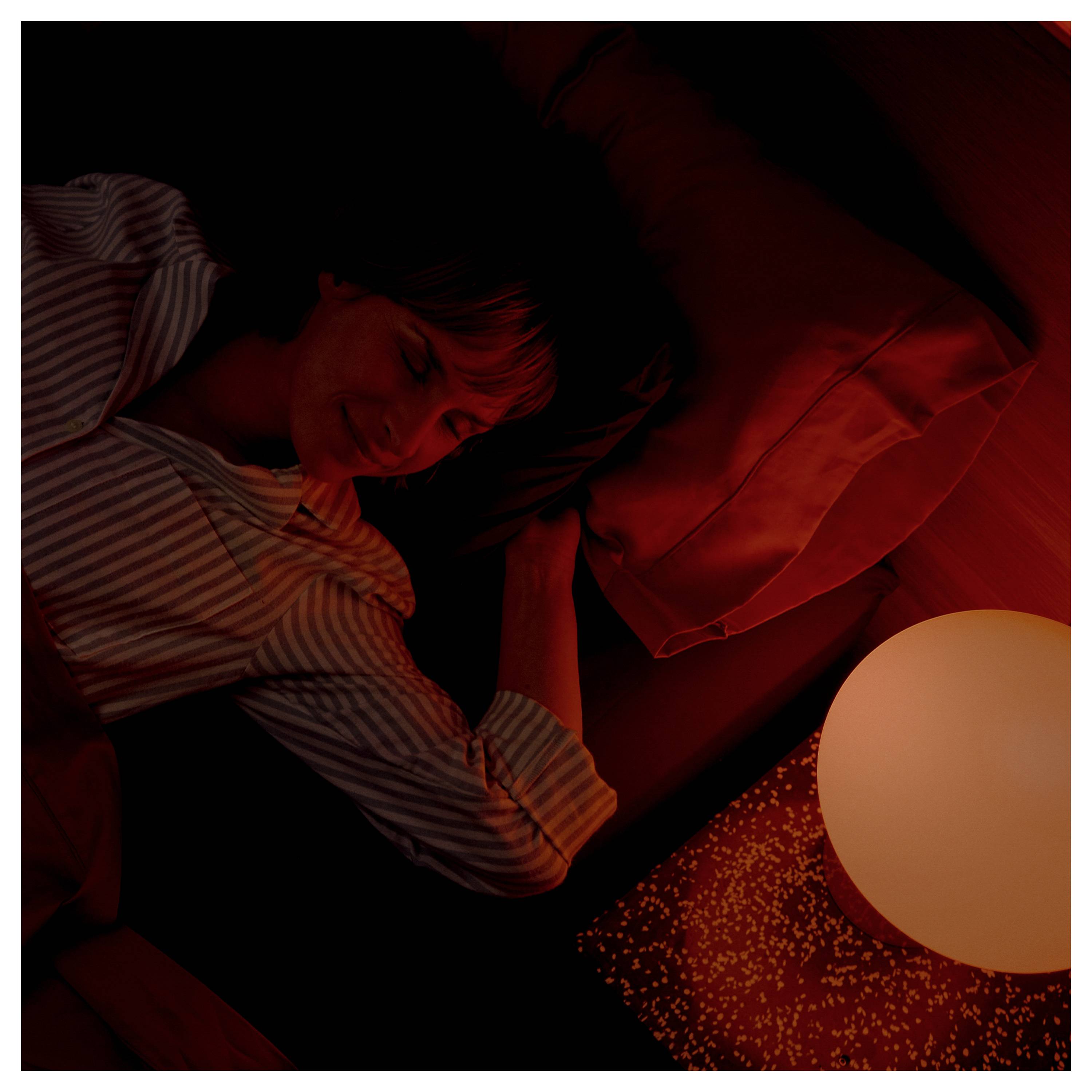 Eine Person schläft friedlich in gedämpftem rotem Licht neben einer leuchtenden, kugelförmigen Lampe und erzeugt eine ruhige Nachtatmosphäre.