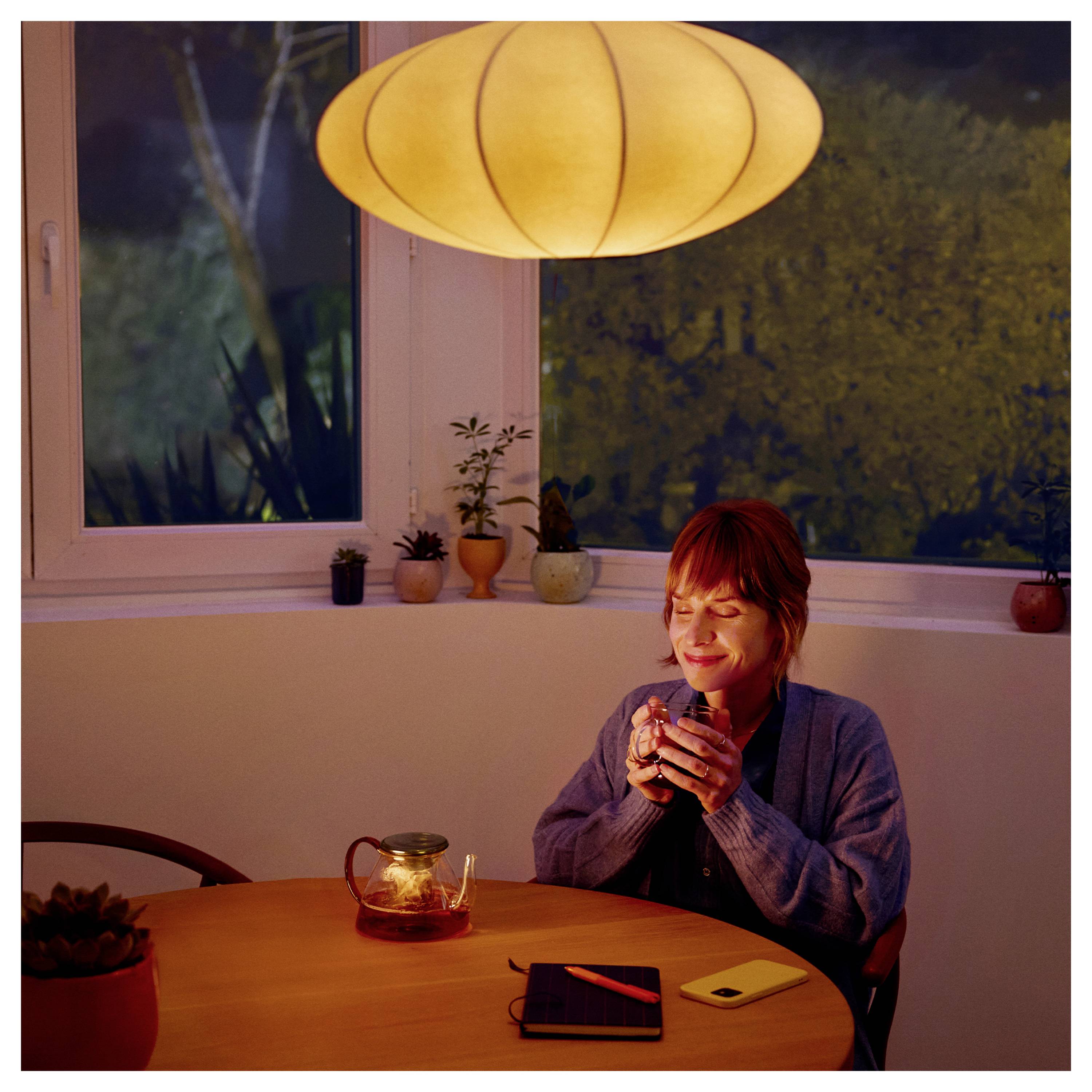 Eine Person, die nachts an einem Tisch sitzt, eine Tasse hält und lächelt. Eine Teekanne und ein Notizbuch befinden sich auf dem Tisch unter einer warmen, glühenden Lampe.