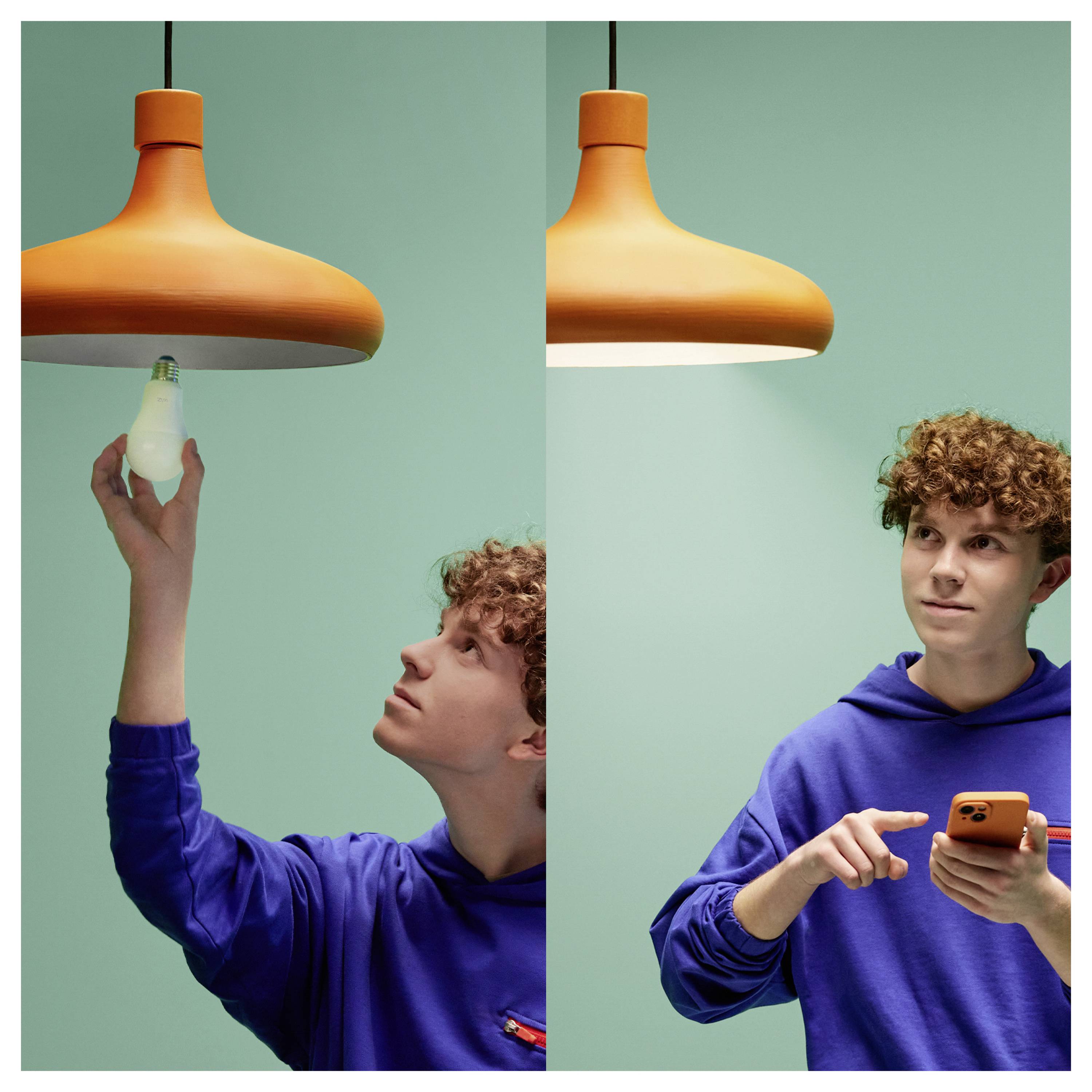 Eine Person in einem blauen Kapuzenpullover wechselt eine Glühbirne in einer orangefarbenen Deckenleuchte und benutzt anschließend ein Smartphone, möglicherweise um Einstellungen anzupassen.