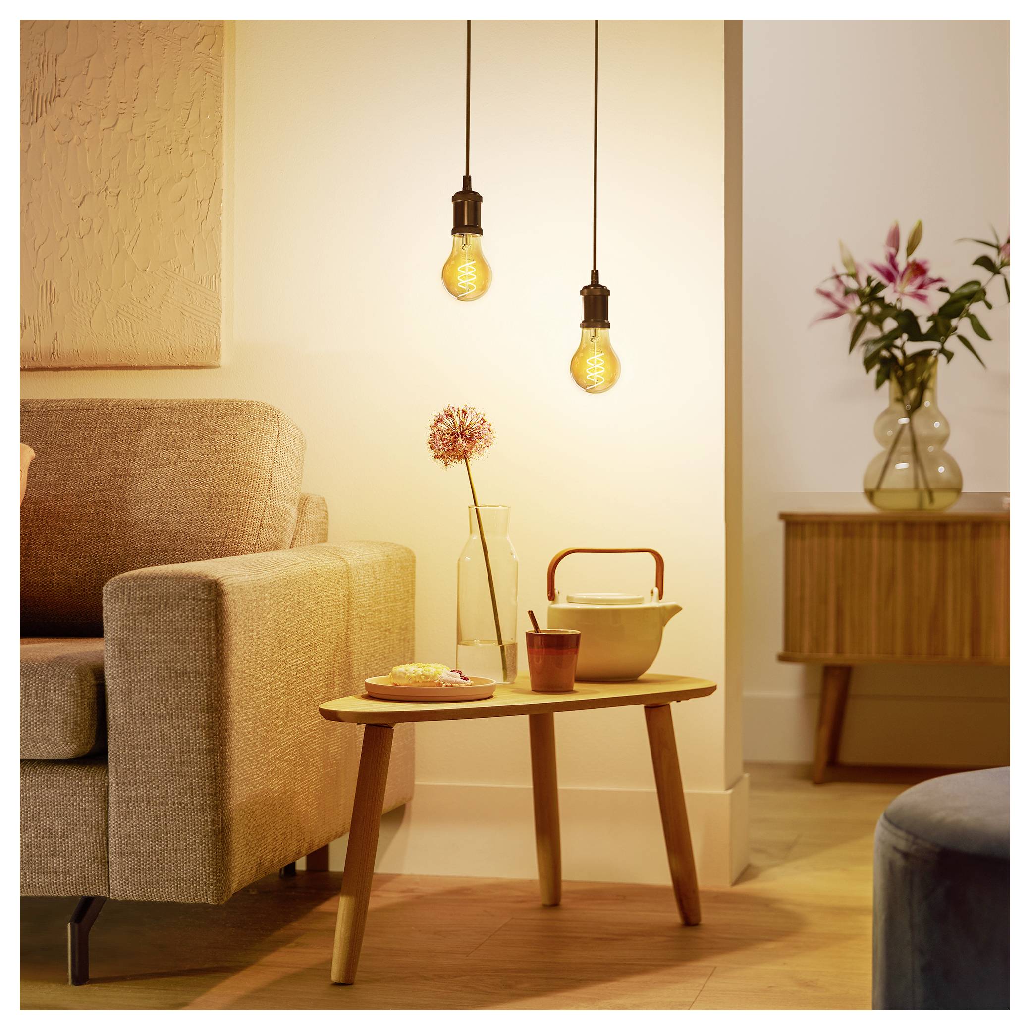 Ein gemütliches Wohnzimmer mit warmem Licht, das ein beige Sofa, einen Holzkaffeetisch mit Teekanne und Tasse sowie zwei hängende Glühbirnen zeigt.