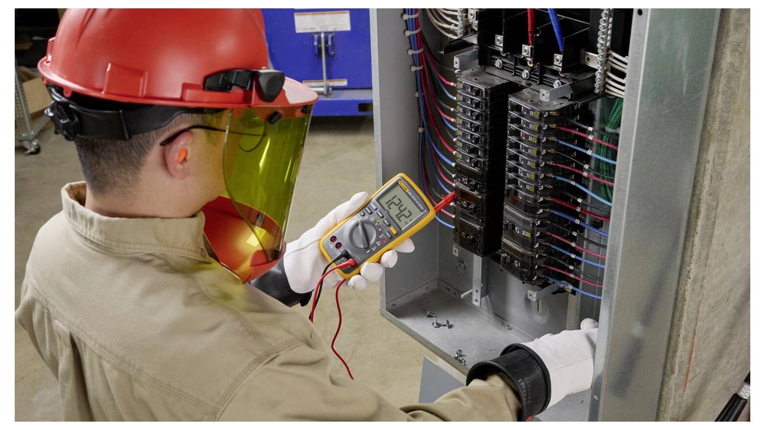 Ein Arbeiter in Schutzausrüstung verwendet ein Multimeter, um die elektrische Verkabelung in einem offenen Schaltschrank zu überprüfen, was eine Sicherheitsinspektionsmaßnahme darstellt.