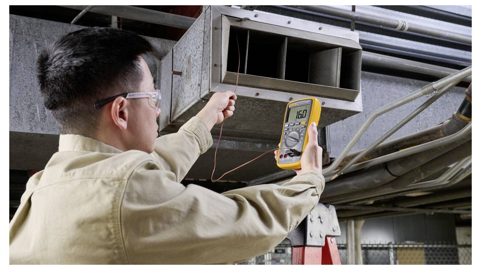 Eine Person, die mit einem digitalen Multimeter in einer industriellen Umgebung nahe einem Luftkanal die Spannung misst und dabei Arbeiten zur elektrischen Überprüfung durchführt.