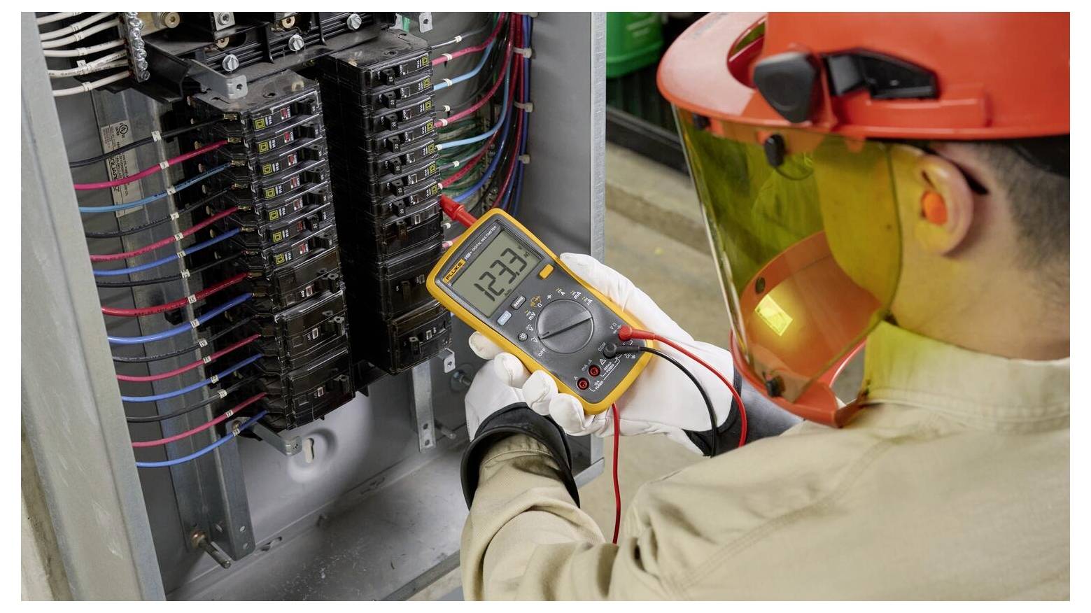 Ein Techniker in Schutzausrüstung verwendet ein Multimeter, um die Spannung in einer elektrischen Verteilerdose zu überprüfen, wobei auf dem Display '123' angezeigt wird.