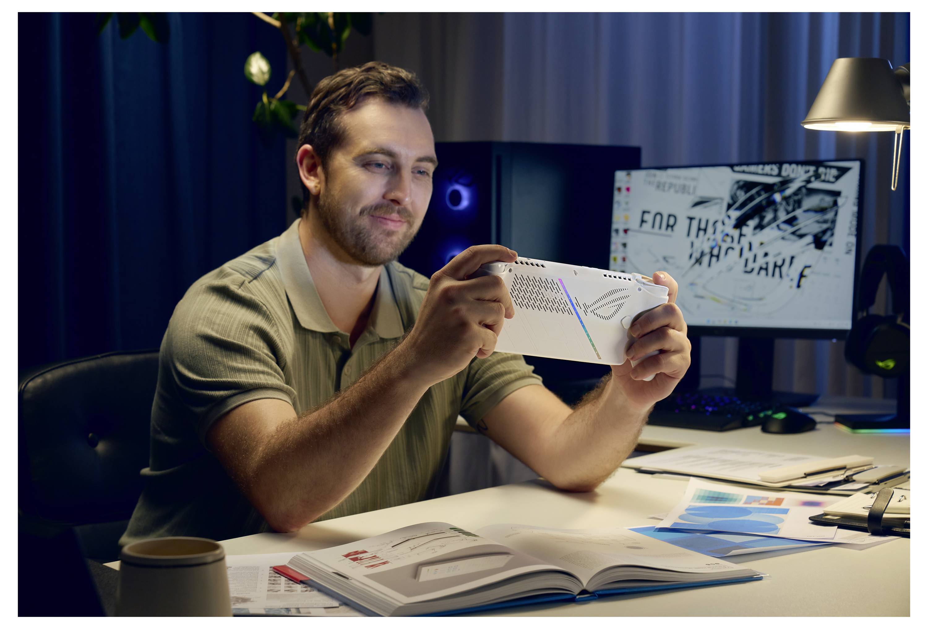 Eine Person sitzt an einem Schreibtisch und lächelt auf ein Gerät. Hinter ihr befindet sich ein beleuchteter Computerbildschirm mit undefinierbarem Inhalt in einer Büroumgebung.