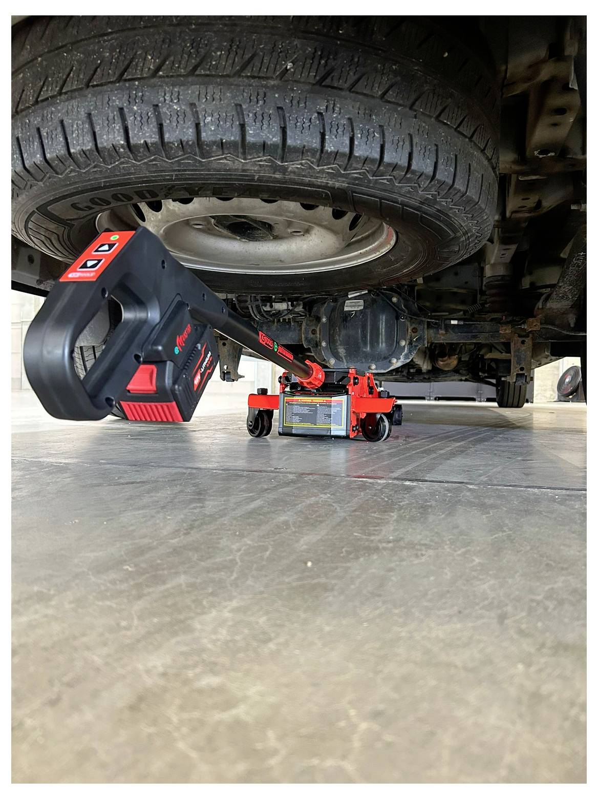 Ein Autoreifen, der von einem hydraulischen Wagenheber angehoben wird, wobei der Hebel und das Bedienfeld des Wagenhebers im Vordergrund sichtbar sind, auf einer Betonoberfläche.