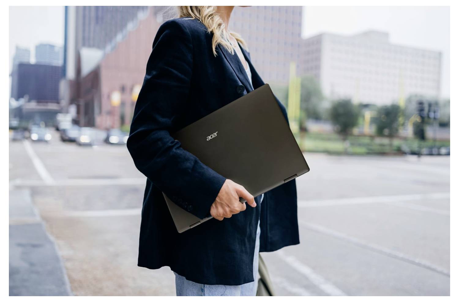 Eine Person in einem dunklen Blazer hält ein Acer-Laptop, während sie auf einer Stadtstraße steht, mit Gebäuden und verschwommenem Verkehr im Hintergrund.