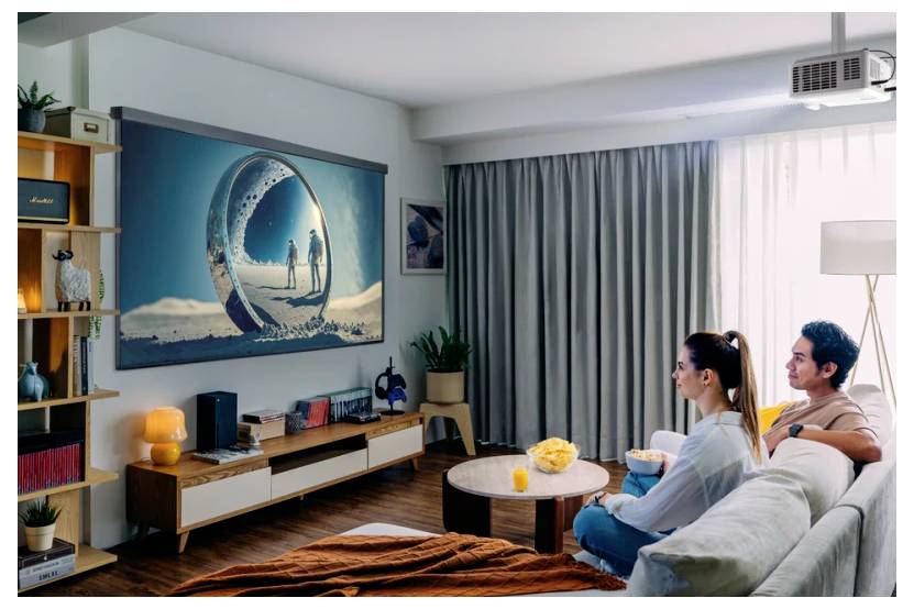 Ein Mann und eine Frau sitzen auf einem Sofa und betrachten eine Projektionsleinwand, die ein Bild von zwei Menschen zeigt, die auf einer sandigen Oberfläche gehen, umrahmt von einem kreisförmigen Rahmen.