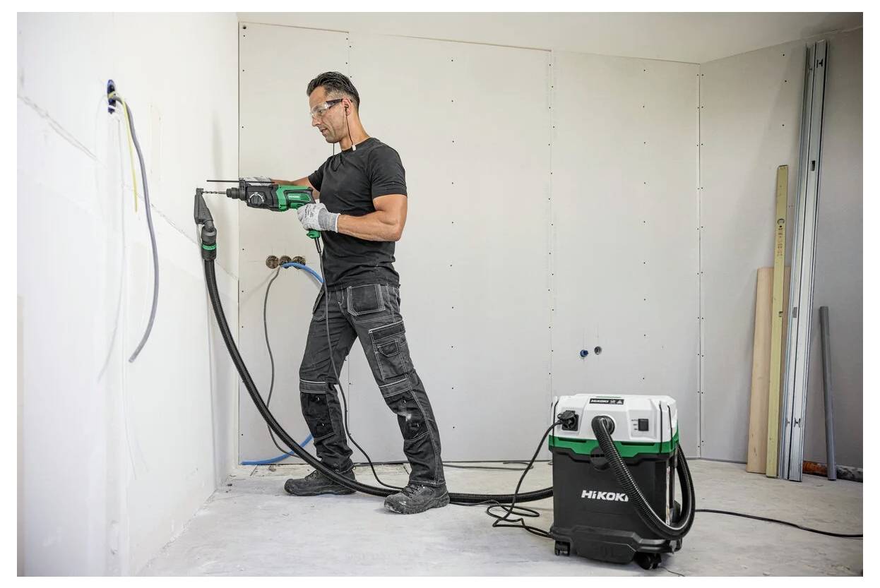 Eine Person in Schutzausrüstung bedient ein Elektrowerkzeug, um in einer teilweise fertiggestellten Wand zu bohren. Ein Staubsauger sammelt in der Nähe Staub.