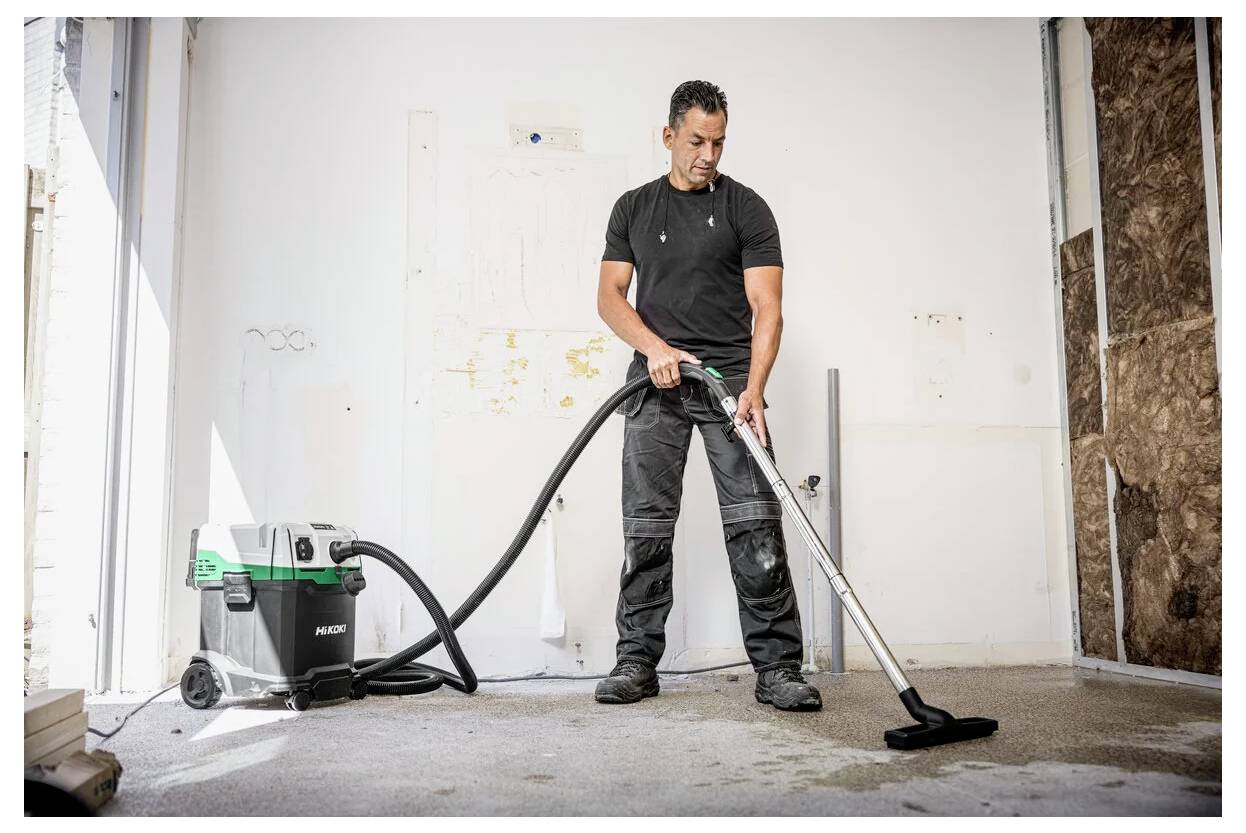 Eine Person saugt einen Boden in einem teilweise renovierten Raum mit einem industriellen Staubsauger und trägt dabei lässige Arbeitskleidung.