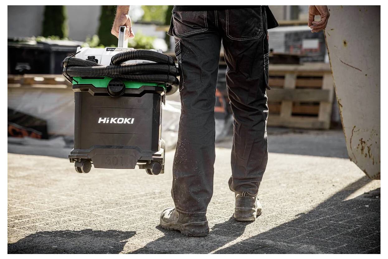 Eine Person trägt einen Hitachi-Werkzeugkoffer auf Rollen und geht über eine sonnenbeschienene Gehsteig. Das Bild vermittelt Mobilität und Arbeitsbereitschaft.