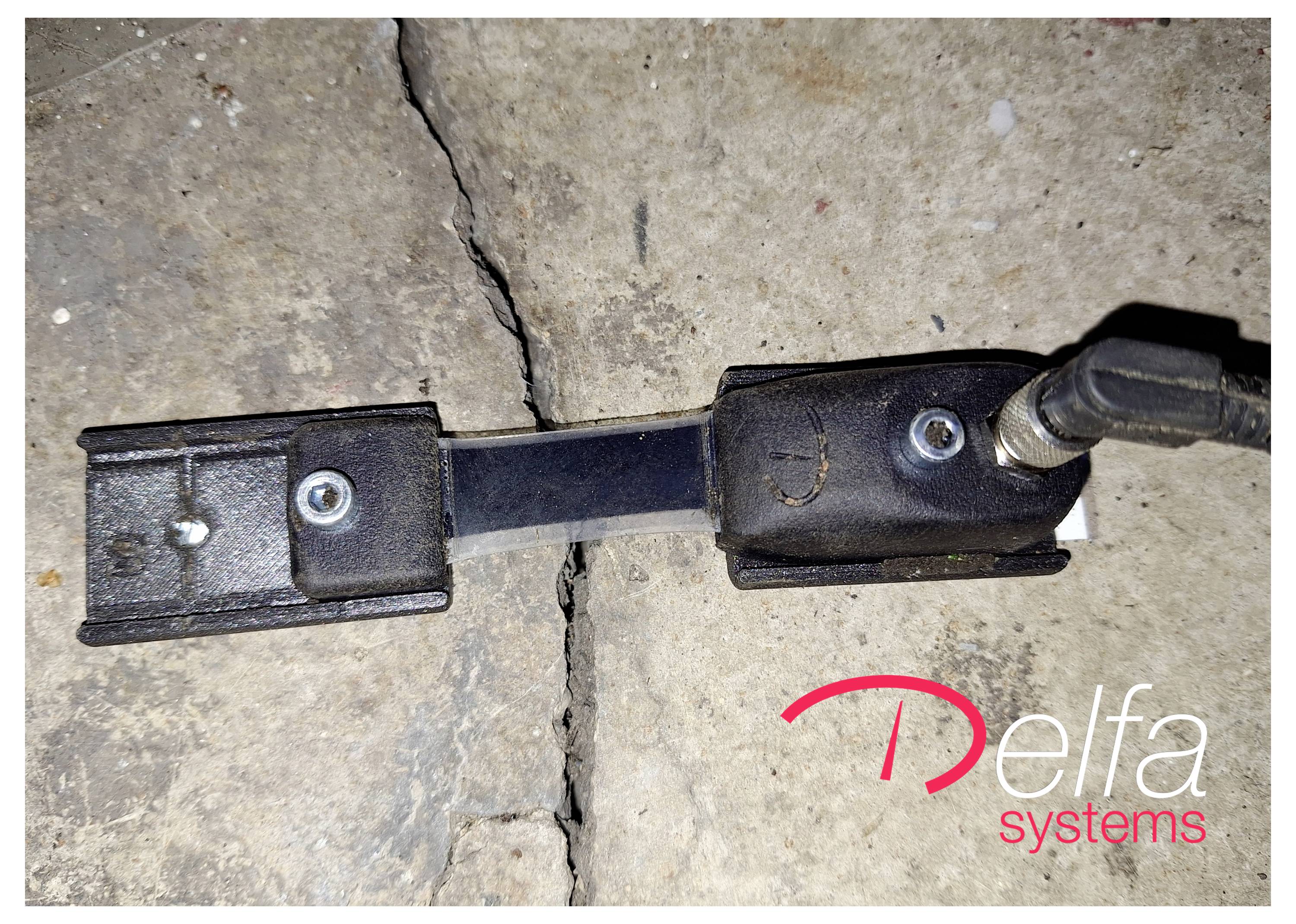 Ein elektronischer Sensor, der auf einem Riss in einer Betonoberfläche montiert ist, mit dem Logo von ‚Delfa Systems' in der unteren rechten Ecke.