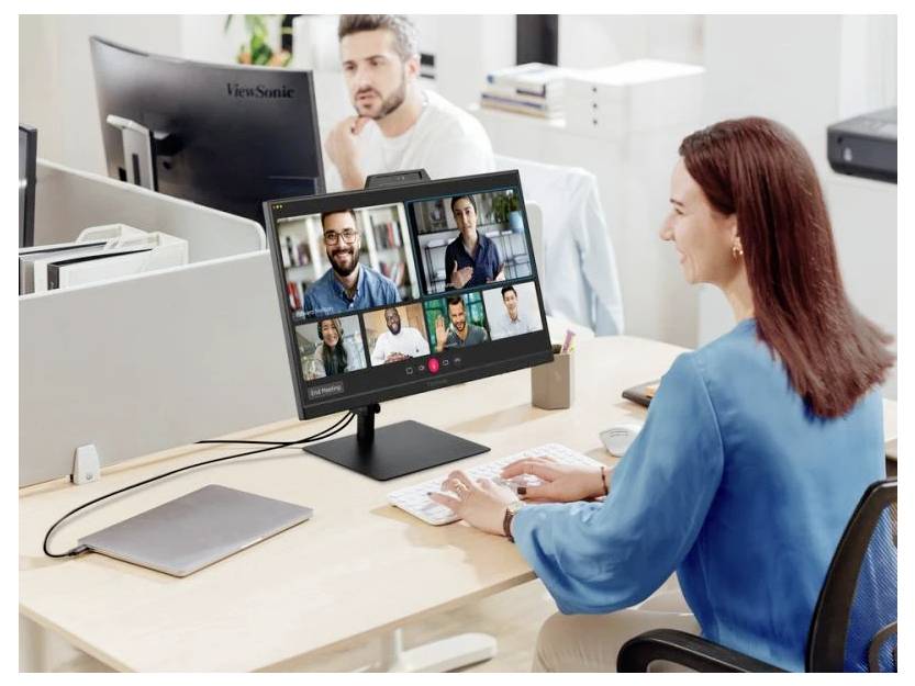 Eine Frau an einem Schreibtisch nimmt an einer Videokonferenz auf ihrem Computerbildschirm teil, auf dem mehrere Teilnehmer zu sehen sind. Ein offener Laptop liegt in der Nähe.