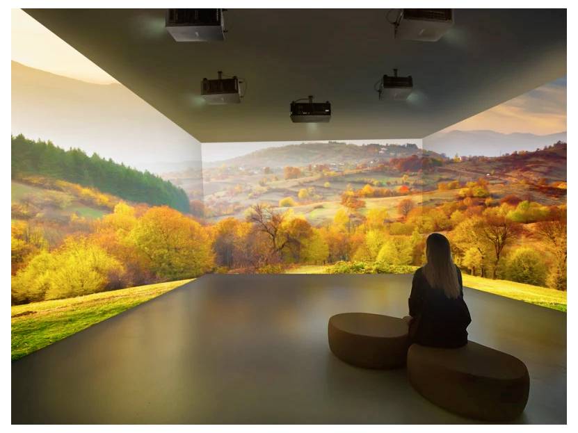 Eine Person sitzt auf einer Bank in einem Raum mit immersiven Bildschirmen, die eine Herbstlandschaft zeigen, mit leuchtend bunten Bäumen und sanft hügeligen Hängen.