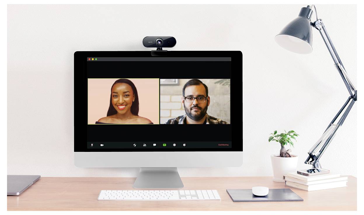 Ein Computerbildschirm, der einen Videoanruf mit zwei Teilnehmern zeigt; eine Person links und eine andere rechts, auf einem Schreibtisch mit Büchern und einer Lampe.