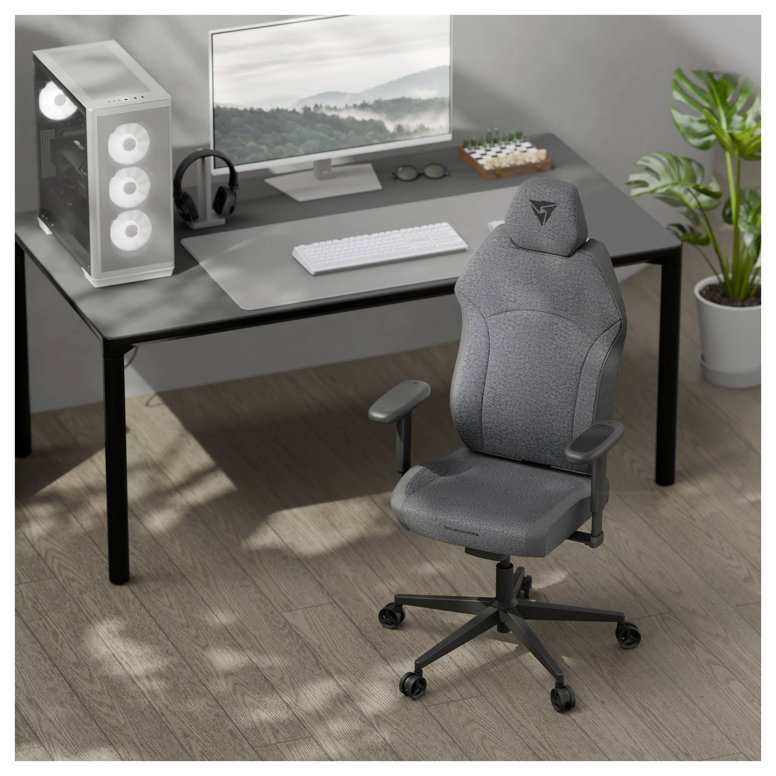 Eine moderne Büroeinrichtung mit einem grauen Gaming-Stuhl auf Rollen, einem schlanken schwarzen Schreibtisch mit einem Monitor, einer Tastatur und einer Topfpflanze daneben.