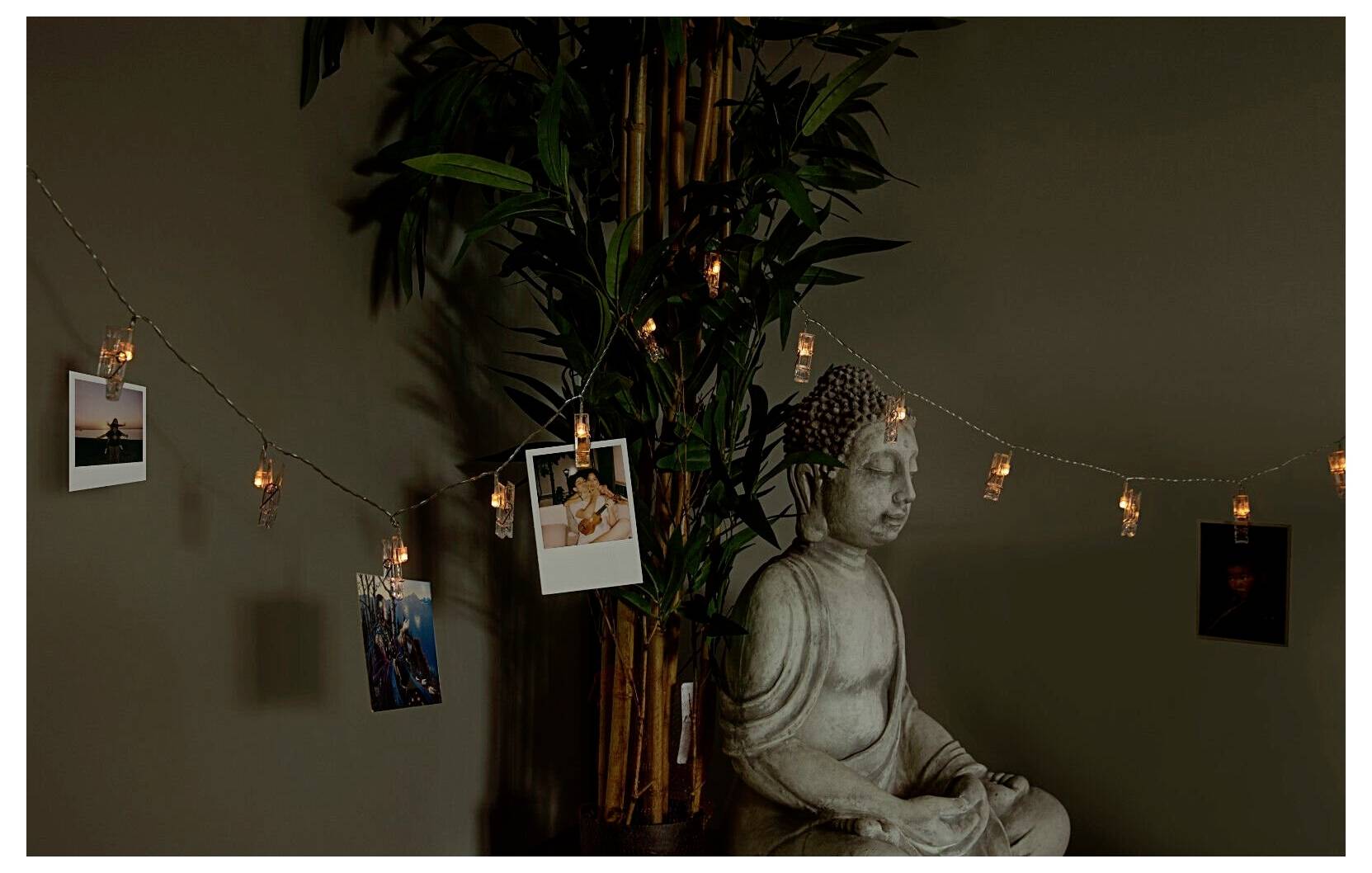 Eine friedvolle Buddha-Statue steht neben einer belaubten Pflanze, mit einer Reihe warmer Lichterketten und Fotos, die eine ruhige Atmosphäre schaffen.