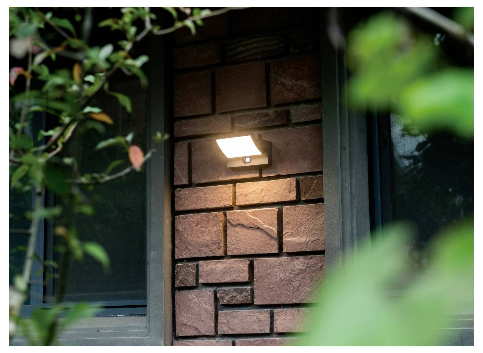 Außenwandleuchte leuchtet an einer Ziegelwand, umgeben von Grün, neben einem Fenster und schafft eine gemütliche Abendatmosphäre.