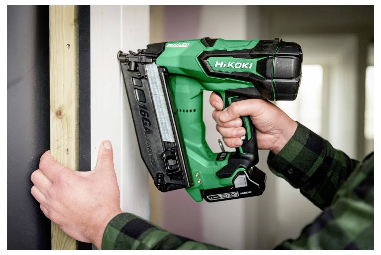 Eine Person verwendet eine grün-schwarze Nagelpistole, um ein Holzbrett an eine Wand zu befestigen. Das Werkzeug ist mit der Marke 'HiKOKI' versehen und verfügt über ein Magazin.