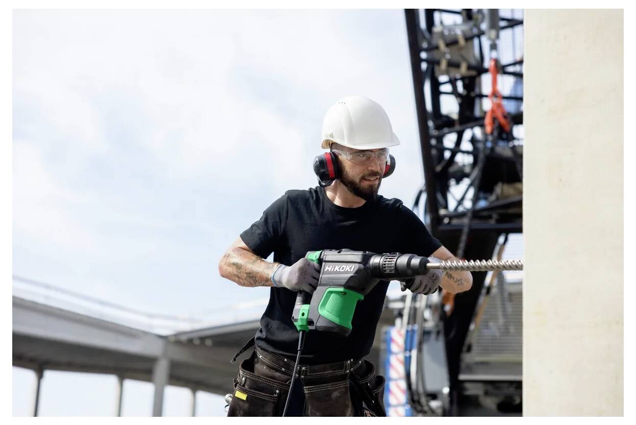 Bauarbeiter mit Sicherheitsausrüstung, der eine Bohrmaschine an einer Betonkonstruktion auf einer Baustelle verwendet. Im Hintergrund ist Baumaschinery zu sehen.