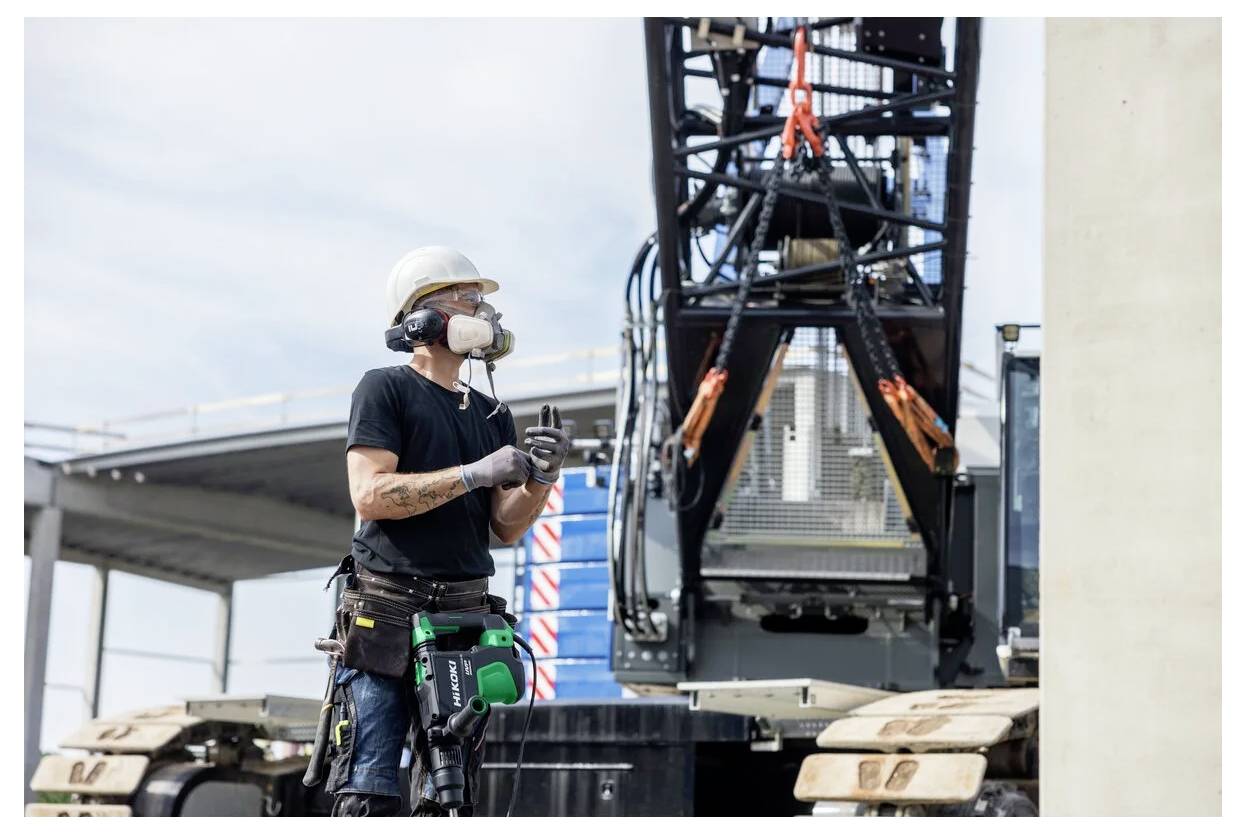 Ein Bauarbeiter mit Schutzhelm und Sicherheitsmaske steht in der Nähe eines großen Stück schwerer Baumaschine auf einer Baustelle.