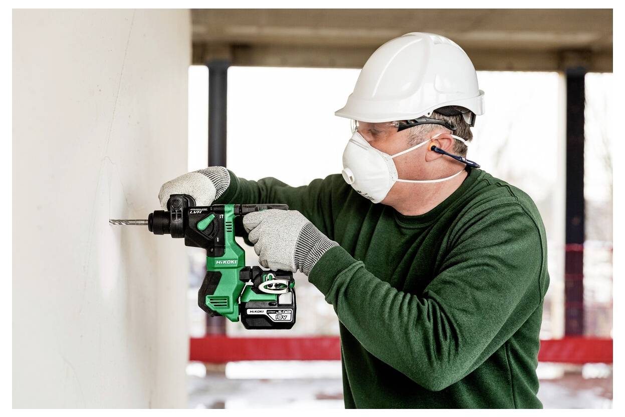 Ein Bauarbeiter mit Sicherheitsausrüstung verwendet einen grünen Bohrer an einer Wand. Der Arbeiter trägt einen Schutzhelm, eine Maske, Handschuhe und Schutzbrille.