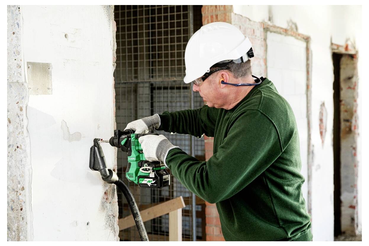 Ein Bauarbeiter mit weißem Schutzhelm und grünem Hemd bohrt mit einer Bohrmaschine an einer Wand in einem Gebäude, das renoviert wird.