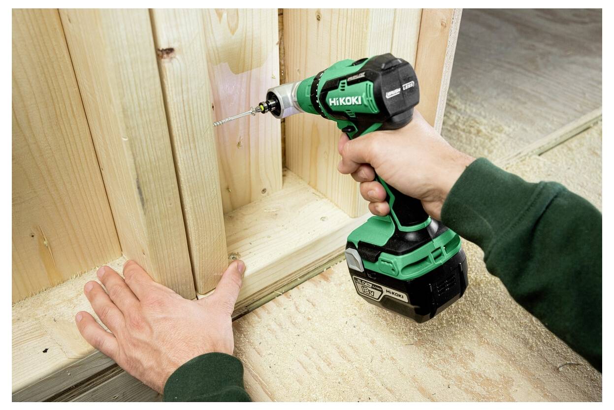 Eine Person benutzt einen grün-schwarzen elektrischen Bohrer, um ein Loch in ein Holzbrett zu bohren, wobei eine Hand das Brett ruhig hält.