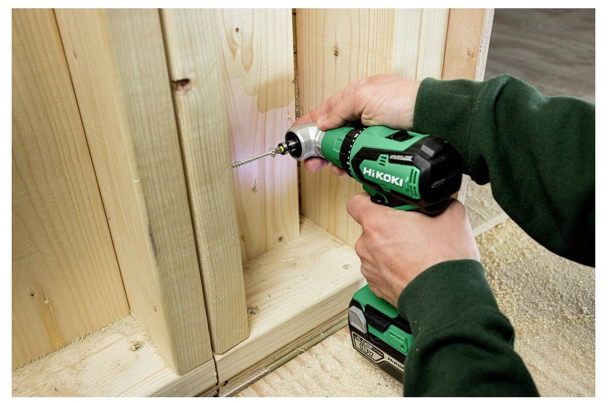 Eine Person verwendet eine grüne kabellose Bohrmaschine, um eine Schraube in Holzbretter zu treiben. Die Szene deutet auf Bauarbeiten oder Holzhandwerksarbeiten hin.