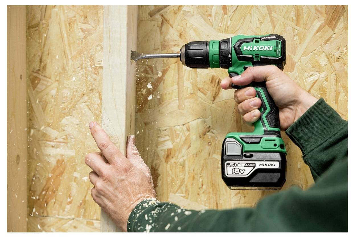 Eine Person verwendet eine grüne kabellose Bohrmaschine mit aufgesetztem Bohrer, um ein Loch in einen vertikalen Holzbalken zu bohren, umgeben von Sperrholzplatten.