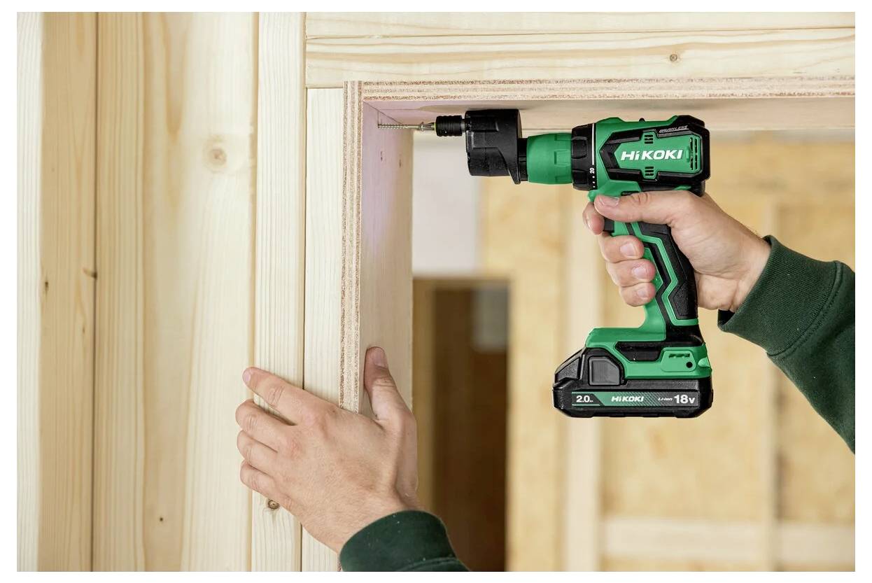 Eine Person verwendet eine grüne kabellose Bohrmaschine, um einen Holztürrahmen in einem Haus im Bau zu installieren. Die Bohrmaschine ist mit 'HIKOKI' gekennzeichnet.