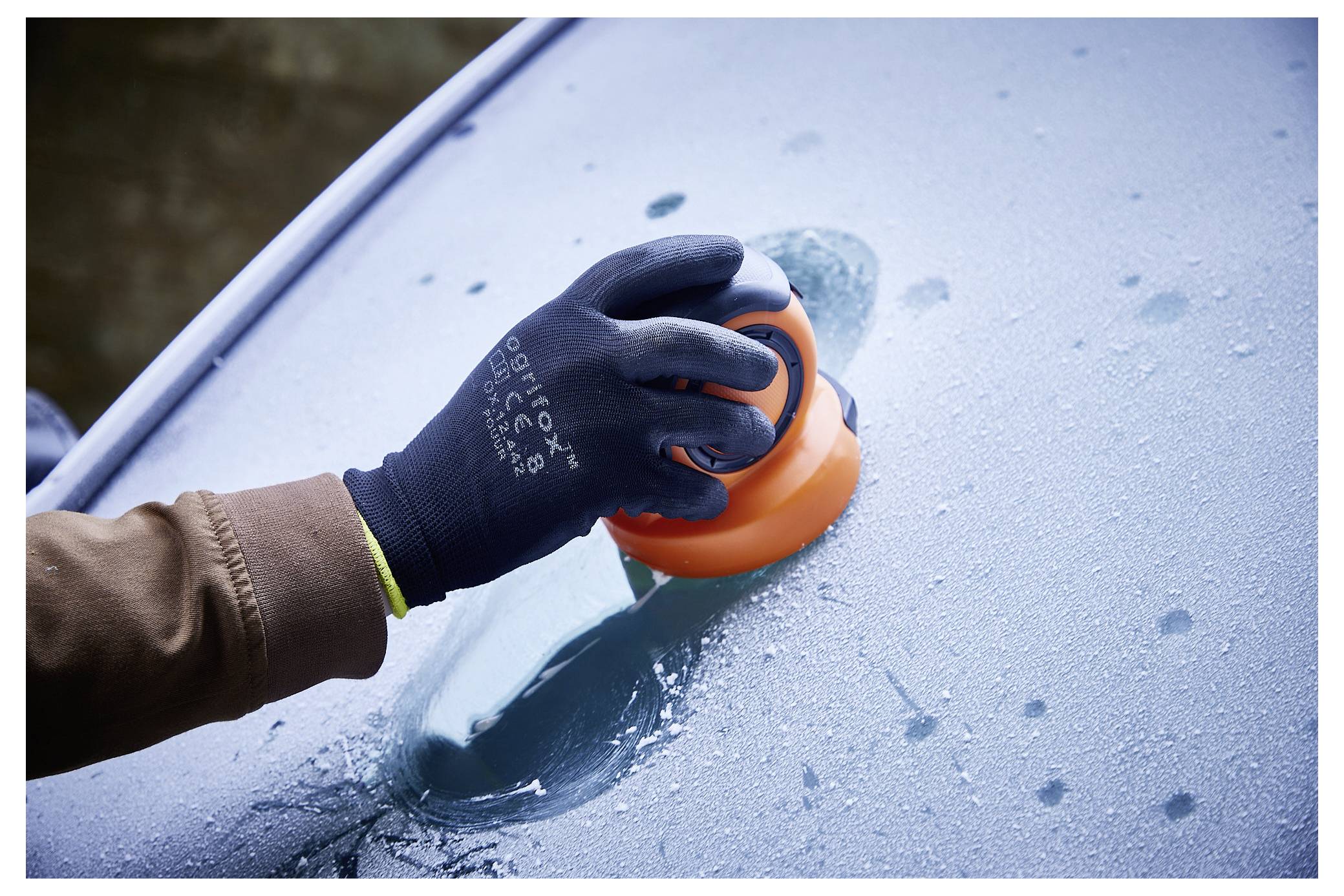 Eine mit einem Handschuh bekleidete Hand, die mit einem orangefarbenen Eiskratzer Frost von einer Autoscheibe befreit.