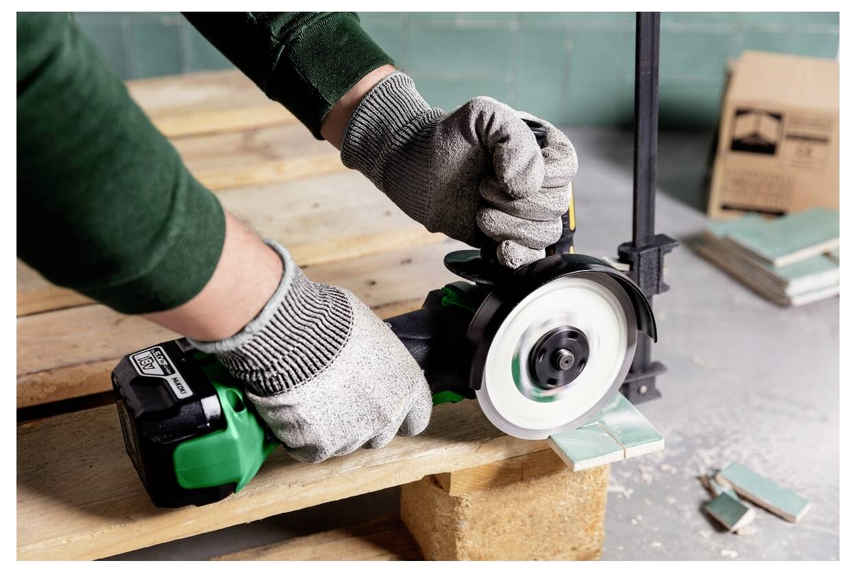 Eine Person mit Handschuhen verwendet ein grünes Elektrowerkzeug, um eine Fliese auf einer Holzpalette zu schneiden, wobei ein Stapel Fliesen und eine Klemme in der Nähe liegen.