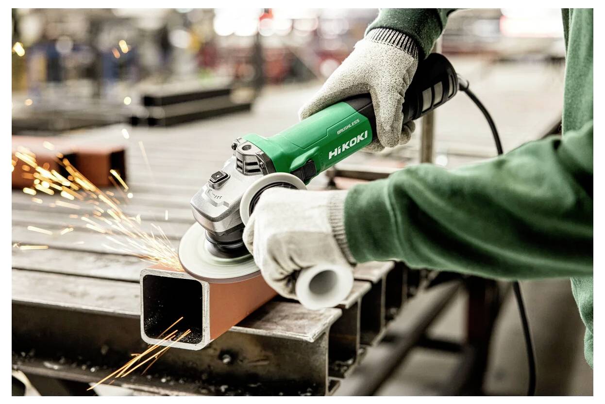 Eine Person mit Handschuhen verwendet eine grüne Hitachi-Powertrennschleifer, um die Kanten eines Metallträgers zu glätten, wobei Funken beim Kontakt aufsprühen.