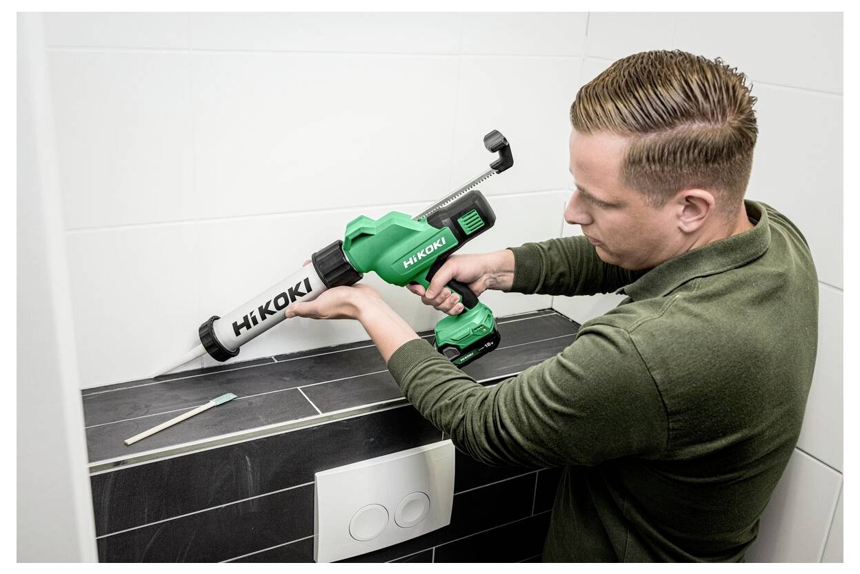 Eine Person trägt Dichtungsmittel auf Badezimmerfliesen auf, indem sie eine grüne Hikoki-Kartuschenpistole verwendet, wobei eine Zahnbürste neben den Fliesen sichtbar ist.