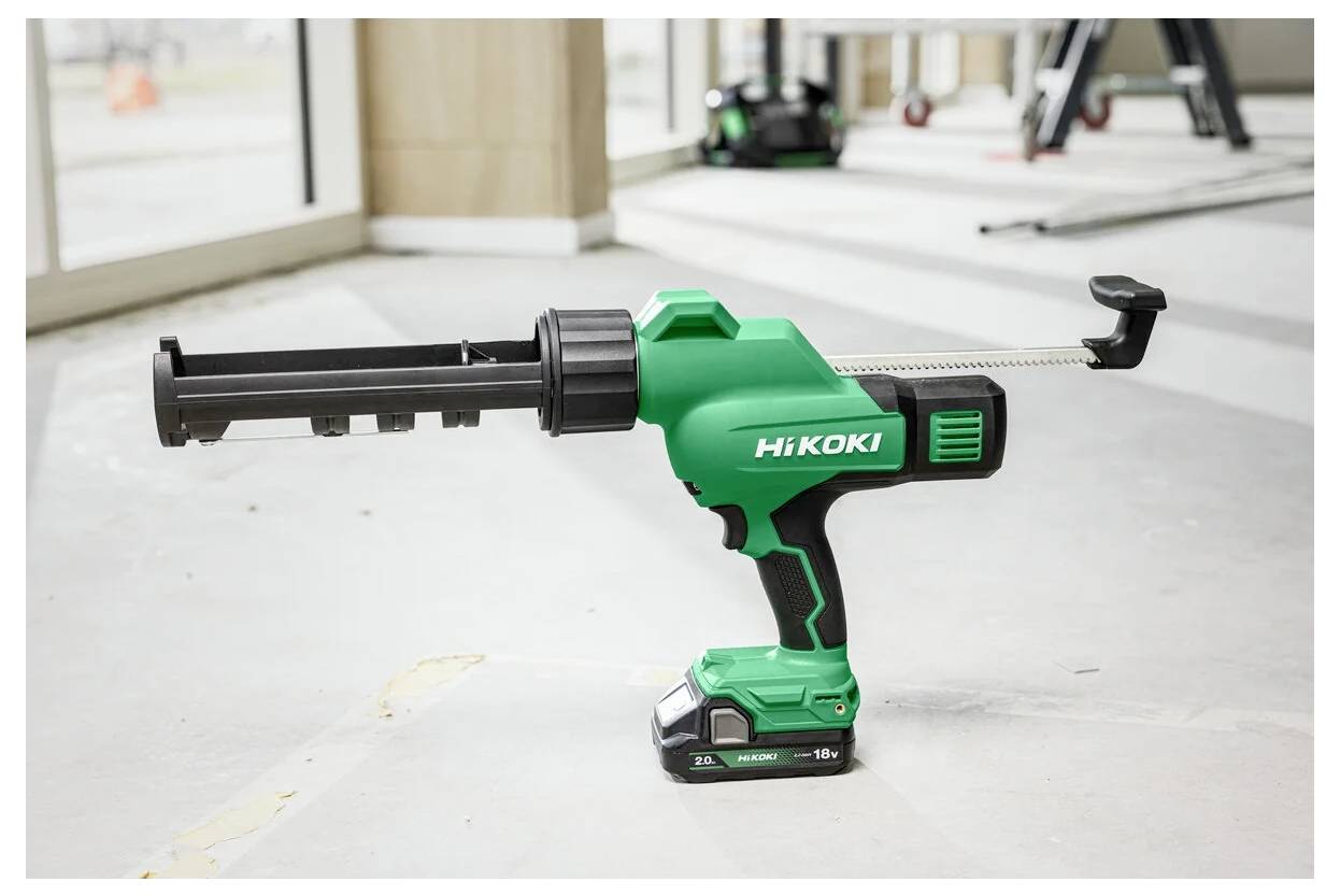 Grüne Hikoki-Akku-Kartuschenpistole auf einem Betonboden in einem hellen Raum, mit großen Fenstern und Bauarbeitsgeräten im Hintergrund.