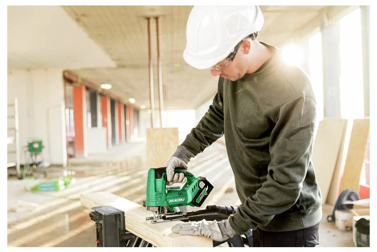 Ein Bauarbeiter mit Schutzhelm und Handschuhen benutzt eine grüne Kreissäge, um Holzbretter in einem teilweise errichteten Gebäude zu schneiden.